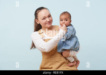 Porträt einer schönen jungen Mutter in beigen Overalls Holding entzückenden kleinen Baby Mädchen in Jeans Kleid in ihre Arme gegen blass blauen Hintergrund. Baby Stockfoto