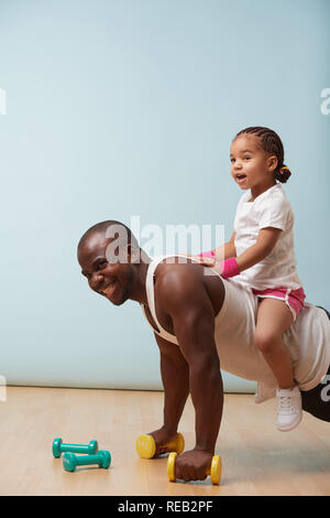 Stattlichen schwarzen Jungen Vater tut, Push-ups auf kurzhanteln mit seinem süßen kleinen Tochter ihn reiten. Sie beide genießen Sie grosse Zeit. Im Innenbereich. Stockfoto