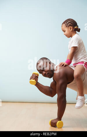 Stattlichen schwarzen Jungen Vater tut, Push-ups auf kurzhanteln mit seinem süßen kleinen Tochter ihn reiten. Im Innenbereich. Er ist ernsthaft Kamera tensin Stockfoto