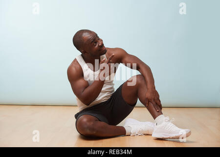 Schönen fetten schwarzen Mann in Weiß ärmelloses Shirt gegen blass blauen Hintergrund. Das Sitzen auf dem Boden, Charmant lächelnd und wies auf die Decke. Stockfoto