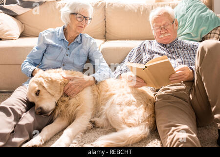 Portrait von Happy senior Paar Kuscheln mit Hund und lesen Bücher auf dem Boden zu Hause sitzen im Sonnenlicht Stockfoto