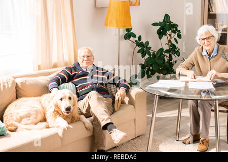 Volle Länge Portrait von gerne ältere Paare zu Hause sowohl auf Kamera und lächelnd im gemütlichen Wohnzimmer Interieur mit niedlichen Hund Stockfoto