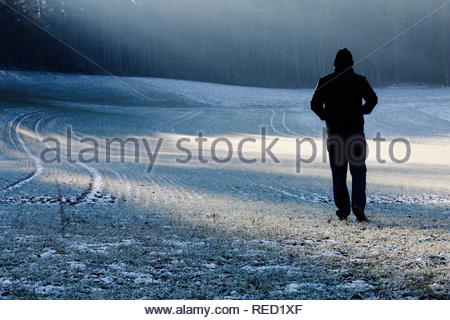 Ein Mann in einer Jacke und Kappe Überqueren einer Frost bedeckt Feld in Bayern an einem Winter. Stockfoto