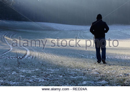 Ein Mann in einer Jacke und Kappe Überqueren einer Frost bedeckt Feld in Bayern an einem Winter. Stockfoto