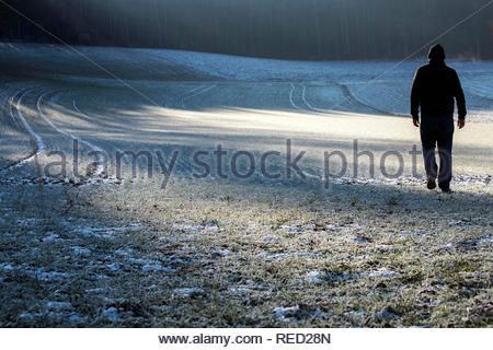 Ein Mann in einer Jacke und Kappe Überqueren einer Frost bedeckt Feld in Bayern an einem Winter. Stockfoto