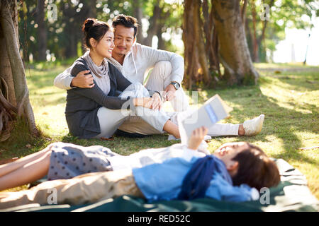 Jungen asiatischen Paar auf Gras im Park chatten mit zwei Kinder lügen Buch lesen im Vordergrund. Stockfoto