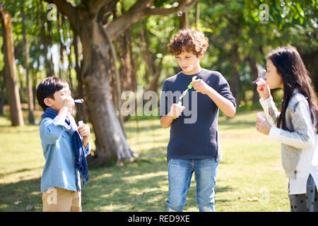 Zwei asiatische und italienische Kinder im Freien spielen im Park Seifenblasen Stockfoto