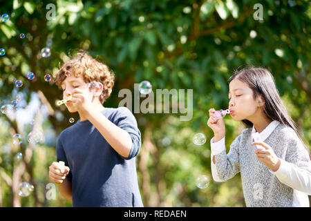 Wenig asiatisches Mädchen und kaukasischen Jungen zusammen spielen bläst Seifenblasen im Freien in einem Park.