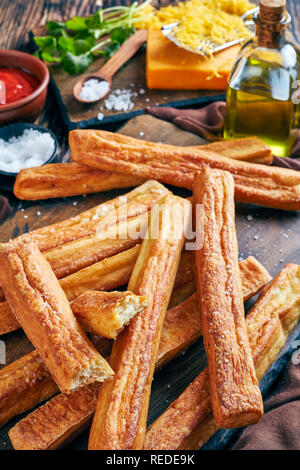 Ansicht von oben von Käse Strohhalme, kitschigen Brot klebt auf einem Holzbrett mit Geriebener Cheddar-Käse, Tomatenmark, Olivenöl auf einem Tisch, vertikal Stockfoto