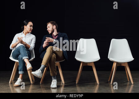 Multiethnischen paar Sitzen auf Stühlen und Reden auf Schwarz isoliert Stockfoto