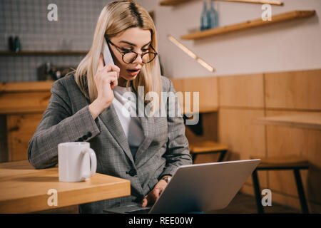 Selektiver Fokus der Schockiert Frau in Gläser sprechen auf dem Smartphone und Laptop suchen Stockfoto