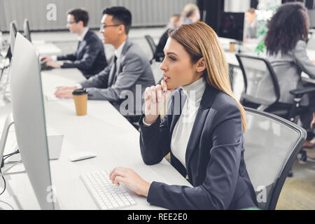 Konzentrierte junge Geschäftsfrau, die mit dem Desktop Computer während der Arbeit mit den Kollegen im Open Space Office Stockfoto
