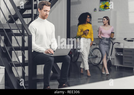 Junger Mann mit Laptop auf Treppen und Unternehmerinnen zu Fuß hinter im Büro ausgerichtet Stockfoto