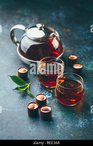 Zwei Glas Tassen und Teekanne mit schwarzem Tee und Pralinen auf dunklem Hintergrund. Stockfoto