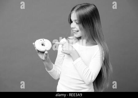 Glückliches Kind zeigen mit dem Finger auf Krapfen auf Violett Hintergrund. Kleines Mädchen mit glasierten ring Donut auf lila Hintergrund. Das ungesunde Essen. Kind Lächeln mit Junk Food. Essen und Nachtisch. Kindheit und Kinderbetreuung. Stockfoto