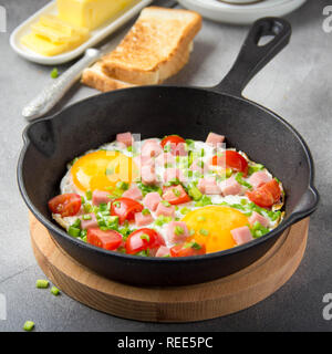 Spiegeleier in Schwarz pan mit Schinken, Tomaten und Frühlingszwiebeln. Köstliches Frühstück mit Toast und Butter. Die klassische Englische Stockfoto