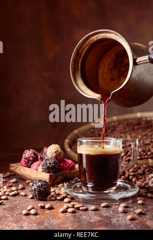 Schwarzen Kaffee in ein kleines Glas Schale von einer Kupfer Kaffeemaschine gegossen. Kaffeebohnen und Pralinen auf einem alten Tisch. Kopieren Sie Platz. Stockfoto