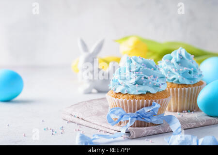 Hausgemachte Vanille Cupcakes mit Streuseln und blauer Schleife dekoriert. Bunny, gefärbte Eier und gelbe Tulpen im Hintergrund. Stockfoto