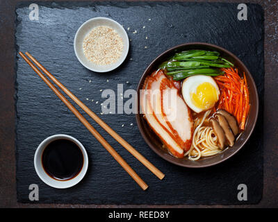 Asiatische Ramen-Suppe mit Huhn, Ei, Karotten, grüne Zwiebeln auf einem dunklen Hintergrund. Ansicht von oben Stockfoto