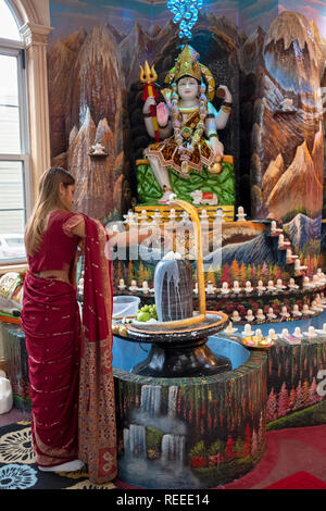 Ein junger Hindu devotee gießt Milch über einen Lingam in der Nähe einer Statue von Shiva an einem Tempel in RichmondHill, Queens, New York City. Stockfoto