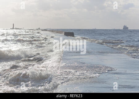 Spritzte Wellen von einem betonpfeiler mit einem Leuchtturm in der Ostsee Hafen mit Frachtschiffe auf dem Raid am Horizont Stockfoto