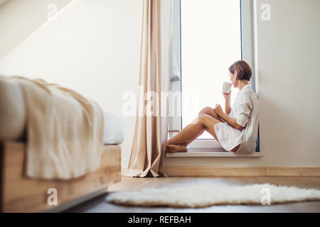 Eine junge Frau mit Nachthemd sitzen durch das Fenster in den Morgen. Stockfoto