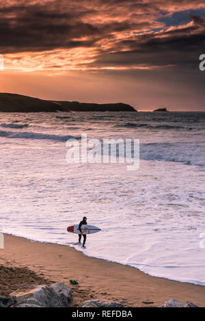 Einen spektakulären Sonnenuntergang als einsame Surfer sein Surfbrett zu Fuß, entlang der Küstenlinie von Fistral Beach in Newquay Cornwall. Stockfoto