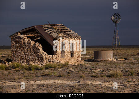 Die untergehende Sonne wirft Licht auf ein verlassenes Haus aus Stein und Windmühle in Eastern New Mexico unter bedrohlichen Himmel Stockfoto