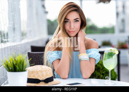 Mädchen in einem Kleid in einer Stadt Cafe sitzen im Sommer. Emotional posiert auf der Suche in den Rahmen. Auf dem Tisch ist ein Glas mit Kalk zu trinken und einen Strohhut. Im Sommer Cafe auf der Veranda entspannen. Stockfoto