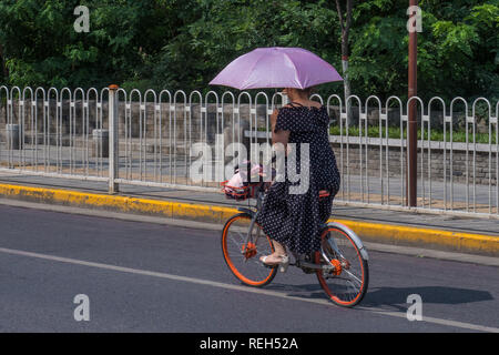 Xian, China, August 2018, Mädchen auf einem Fahrrad mit einem lilafarbenen Sonnenschirm Stockfoto