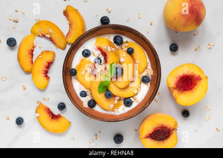 Schüssel Haferflocken mit Joghurt, frische Pfirsiche, Blaubeeren und Minze auf weißem Marmor tisch für gesundes Frühstück. top View Stockfoto