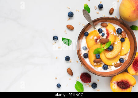 Joghurt mit Pfirsichen, Hafer, Blaubeeren, Honig, Nüsse und Minze in eine Schüssel geben und mit einem Löffel für gesundes Frühstück auf weißem Hintergrund. Ansicht von oben mit Co Stockfoto