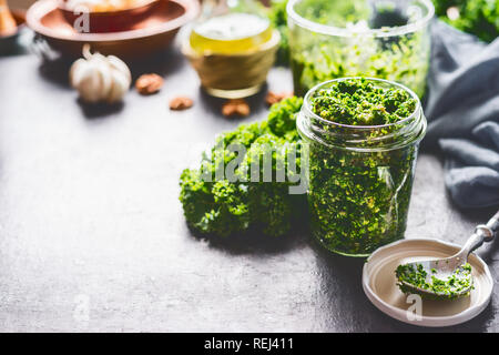 Grünkohl hausgemachtem Pesto Glas stehen auf dunklen rustikalen Küche Tisch Hintergrund mit Zutaten, Ansicht von oben. Kale Vorbereitung. Gesunde detox Gemüse Stockfoto