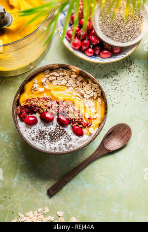 Smoothie Schüssel mit Mango und tropischen Früchten, Chia Samen Joghurt, Pudding und Preiselbeeren, Nüssen, Haferflocken Aufstockung in Kokosnussschalen mit Löffel, oben vie Stockfoto