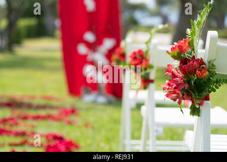 Stühle mit frischen Blumen in roten Tönen für die Trauung eingerichtet auf dem grünen Rasen Stockfoto