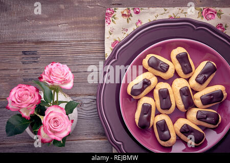 Mini Vanille eclairs mit Schokolade Zuckerguss auf lila Platte, Ansicht von oben auf rustikalen Holztisch mit Rosen und Herzen aus Holz Stockfoto