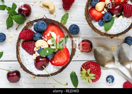 Vegan Kokosmilch Chia Samen Milchreis mit frischen Beeren, Nüsse und Minze in Kokosnuss Schalen mit Löffel über weiße Holztisch. gesundes Frühstück. nach oben v Stockfoto