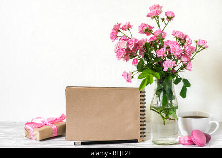 Schönen Blumenstrauß aus Rosen Blumen, Kaffeetasse, Makronen, Geschenkbox und Leere hipster Notebook mit Platz für Text. Urlaub oder Hochzeit Hintergrund Stockfoto