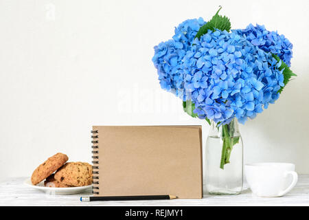Kreative mock up mit einem schönen Blumenstrauß Blaue Hortensie Blumen, Kaffee Tasse, Hafer Cookies und leeren hipster Notebook mit Platz für Text. Holiday o Stockfoto