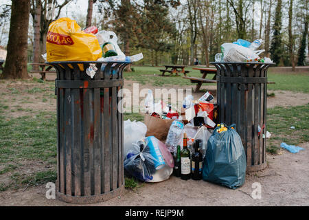 Straßburg, Frankreich - Apr 7, 2018: überquellenden Mülleimer voll mit verschiedenen Arten von Abfall wie Flaschen, pizza Schachteln, Beuteln in der Nähe von einem Picknick ausgewiesenen Bereich mit Holztischen und Bänken in einer französischen Stadt Park an einem frühlingstag ausgestattet. Stockfoto