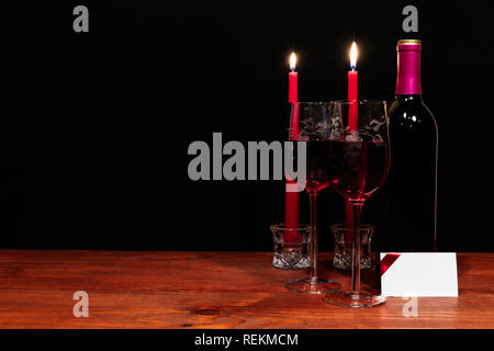 Schöne geätzte Gläser und eine Flasche Rotwein, rote Kerzen, auf Holztisch mit Namensschild auf dunklem Hintergrund. Valentinstag, Muttertag, Ostern, Stockfoto