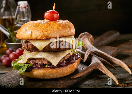 Double Decker wild Burger mit geschmolzenem Käse auf ein knuspriges Brötchen mit Hirschgeweihen neben auf einem rustikalen Tisch oder Theke Stockfoto