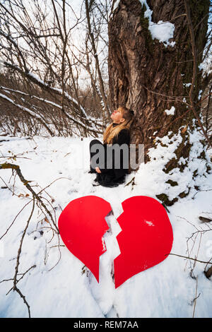 Deprimiert und untröstlich Frau lehnte sich auf Baum im Wald im Winter. Stockfoto