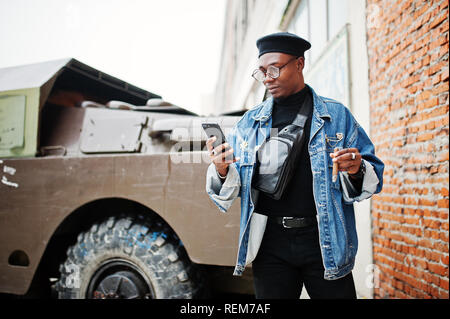 Afrikanische amerikanische Mann in Jeans Jacke, Baskenmütze und Brillen, rauchen Zigarre und stellte gegen btr Limousine, mit Handy zur Hand. Stockfoto