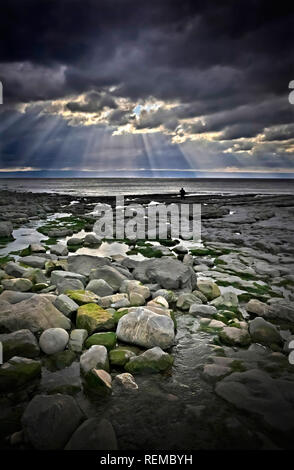 Felsiger Strand mit stürmischem Himmel und dramatischem Licht und eine Person in Silhouette in der Ferne durch Wasser Rand Einsamkeit Konzept Stockfoto