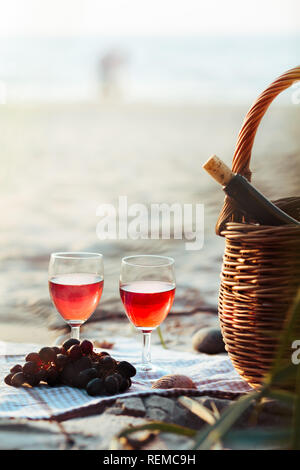 Zwei Weingläser mit Rotwein ständigen Strand, neben Trauben und Weidenkorb mit einer Flasche Wein. Junges Paar im Meer im Hintergrund Stockfoto