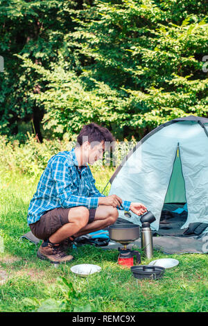 Verbringt einen Urlaub auf dem Campingplatz. Junger Mann Vorbereitung einer Mahlzeit im Freien neben Zelt Stockfoto