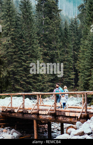 Familie gehen über eine hölzerne Brücke über einem Gebirgsbach. Ausgaben Urlaub auf Wanderschaft mit Rucksäcken in einem Berge und Wälder Stockfoto