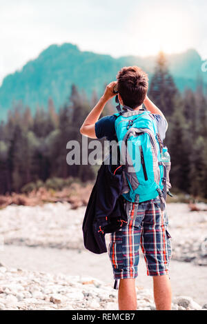 Junge Wanderer mit Rucksack schaut durch ein Fernglas auf Bergspitzen, steht über einem Fluss. Junge verbringt einen Urlaub in den Bergen, Wandern mit Ba Stockfoto