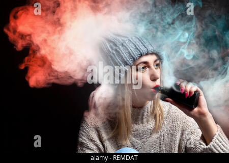Blonde Mädchen in einem gestrickten Pullover und Hut hält eine elektronische Zigarette vape in ihrer Hand mit einer Maniküre und atmet eine Menge in den Farben Rot und Grün smo Stockfoto